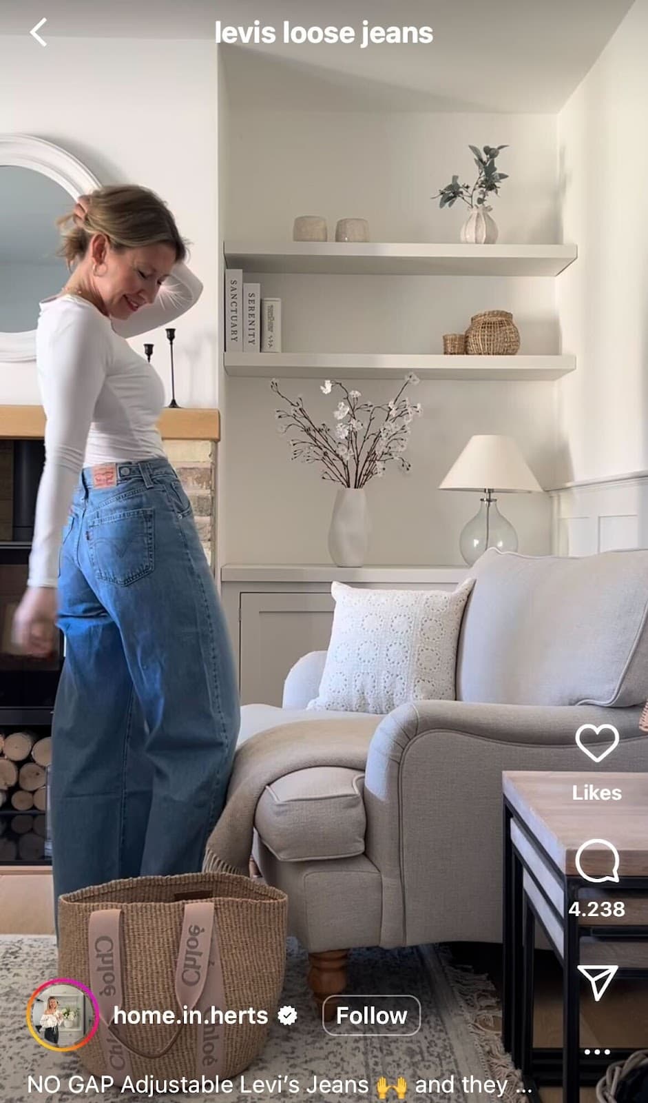 A person with dirty blonde hair, wearing a tight white shirt and baggy jeans, standing in a living room.