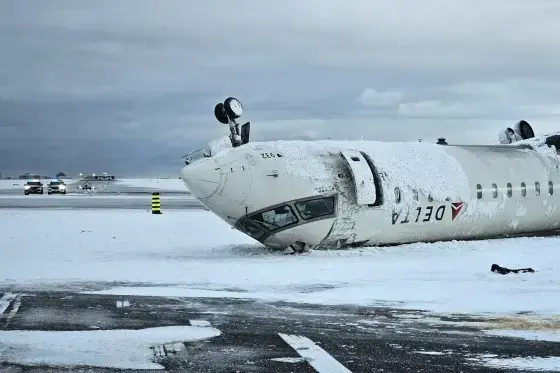 A Delta airplane lies upside down on a snowy runway after a crash, with emergency vehicles in the background.